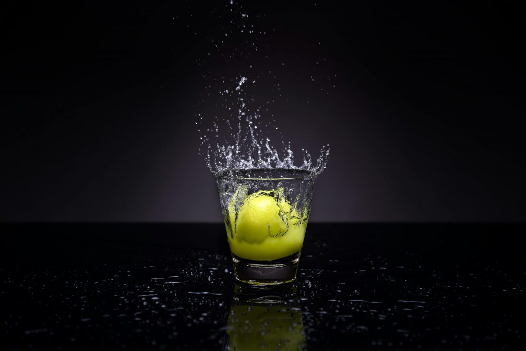A lemon making a splash in a glass of water against a dark background.